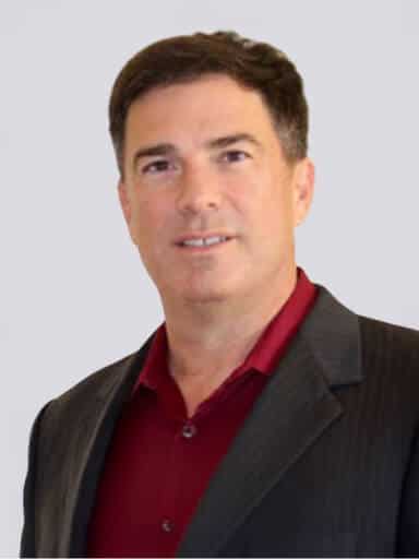 Man wearing a dark suit jacket over a red collared shirt, facing forward against a plain light background.